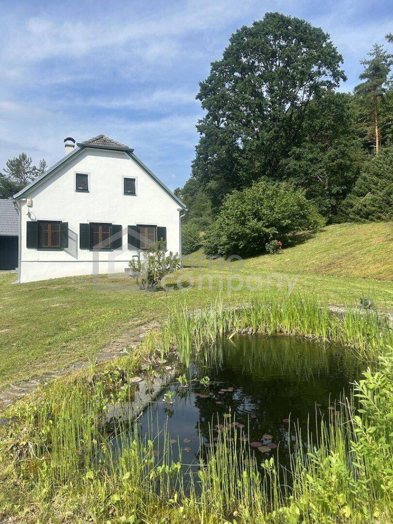 Charmantes Landhaus mit Teich im Vordergrund, umgeben von üppigem Grün und Wald.