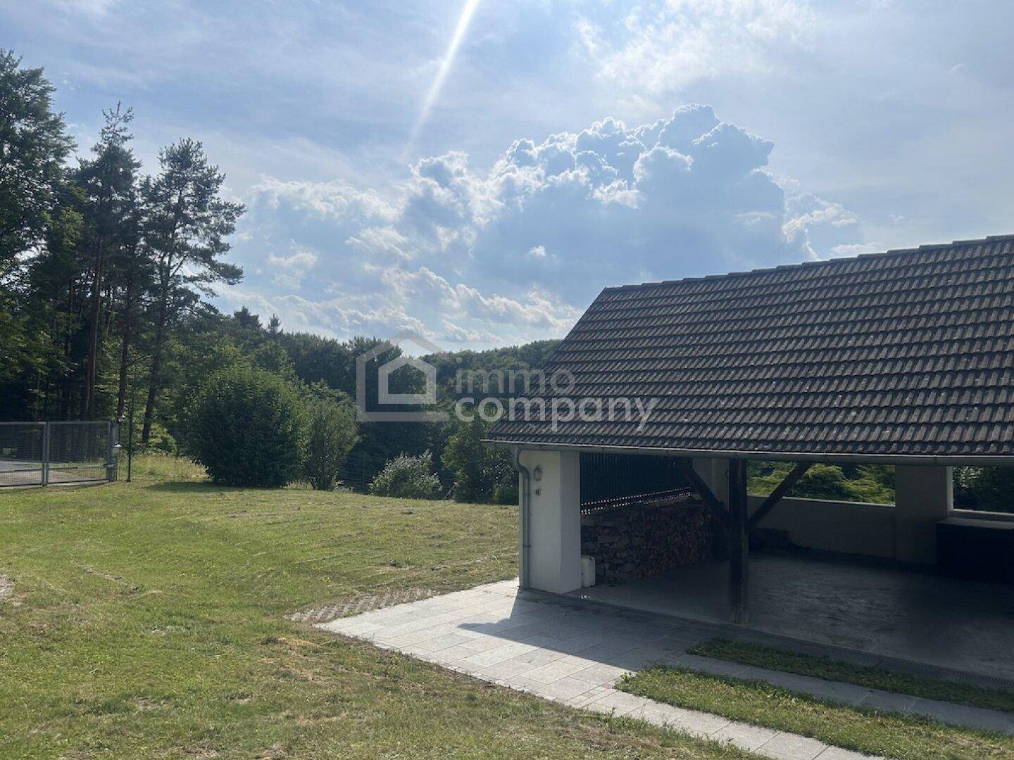 Carport mit Holzstapel und weitem Blick auf die bewaldete Landschaft unter blauem Himmel.