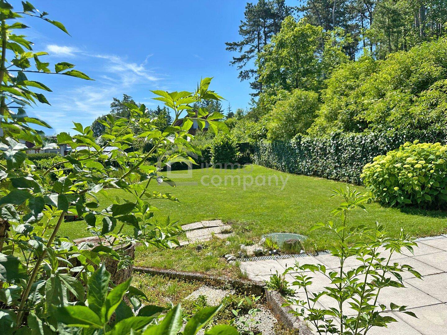 Gepflegte Rasenfläche im Garten mit Steinplatten und üppiger Bepflanzung im Hintergrund.