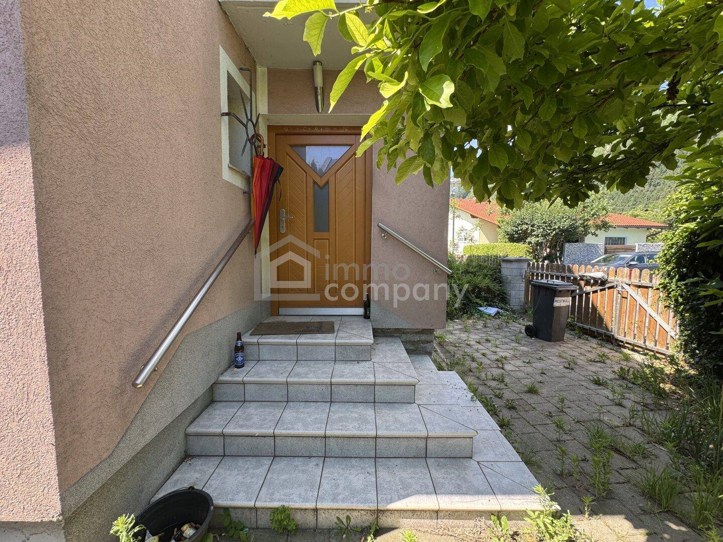 Eingangsbereich des Hauses mit gefliesten Stufen, einem Handlauf und einer Holztür.