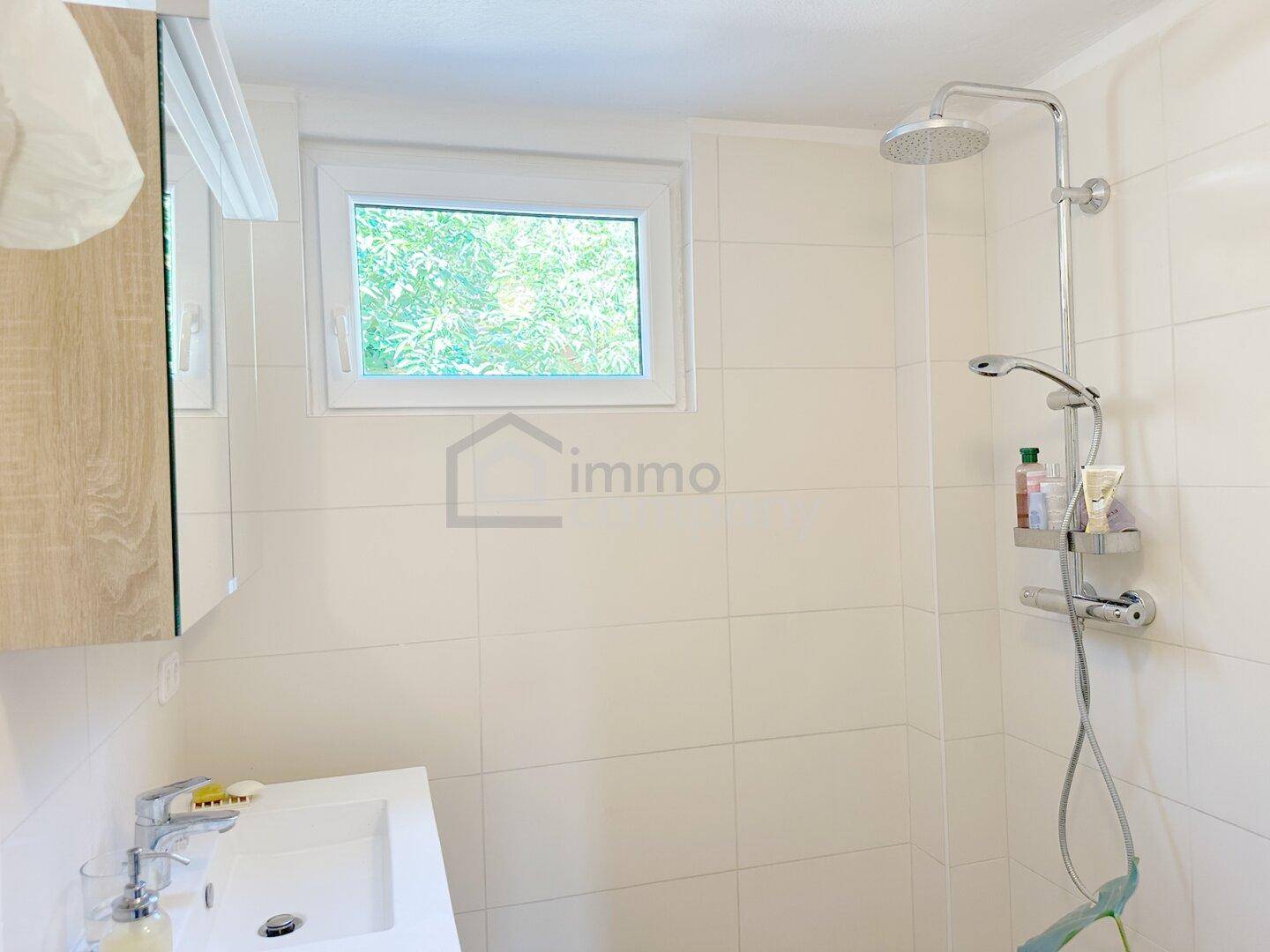 Modernes Badezimmer mit Dusche, Waschbecken und Fenster, hell gefliest in Weiß.