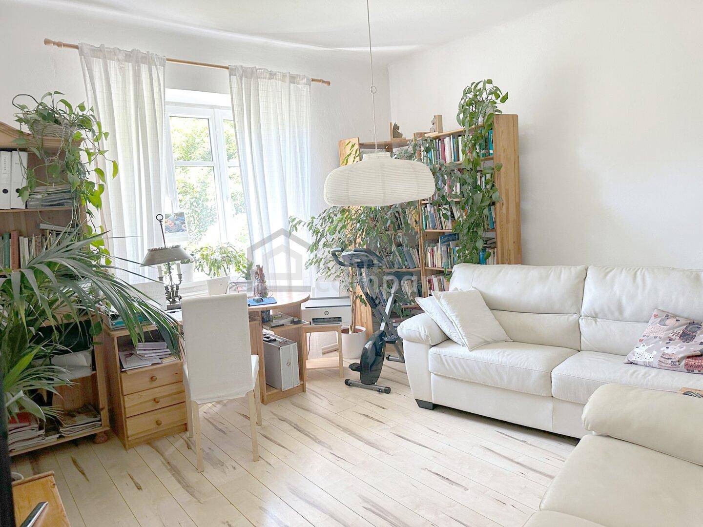 Gemütliches Wohnzimmer mit weißem Sofa, Holzregalen und vielen Pflanzen, hell durch große Fenster.