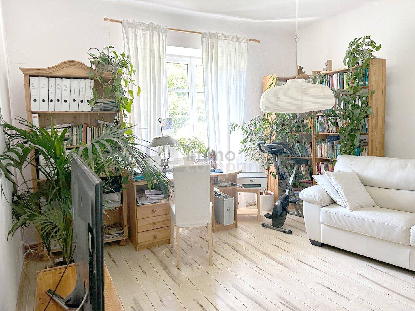 Heller Wohnbereich mit Schreibtisch, weißem Sofa und vielen Pflanzen, ideal für Arbeit und Entspannung.