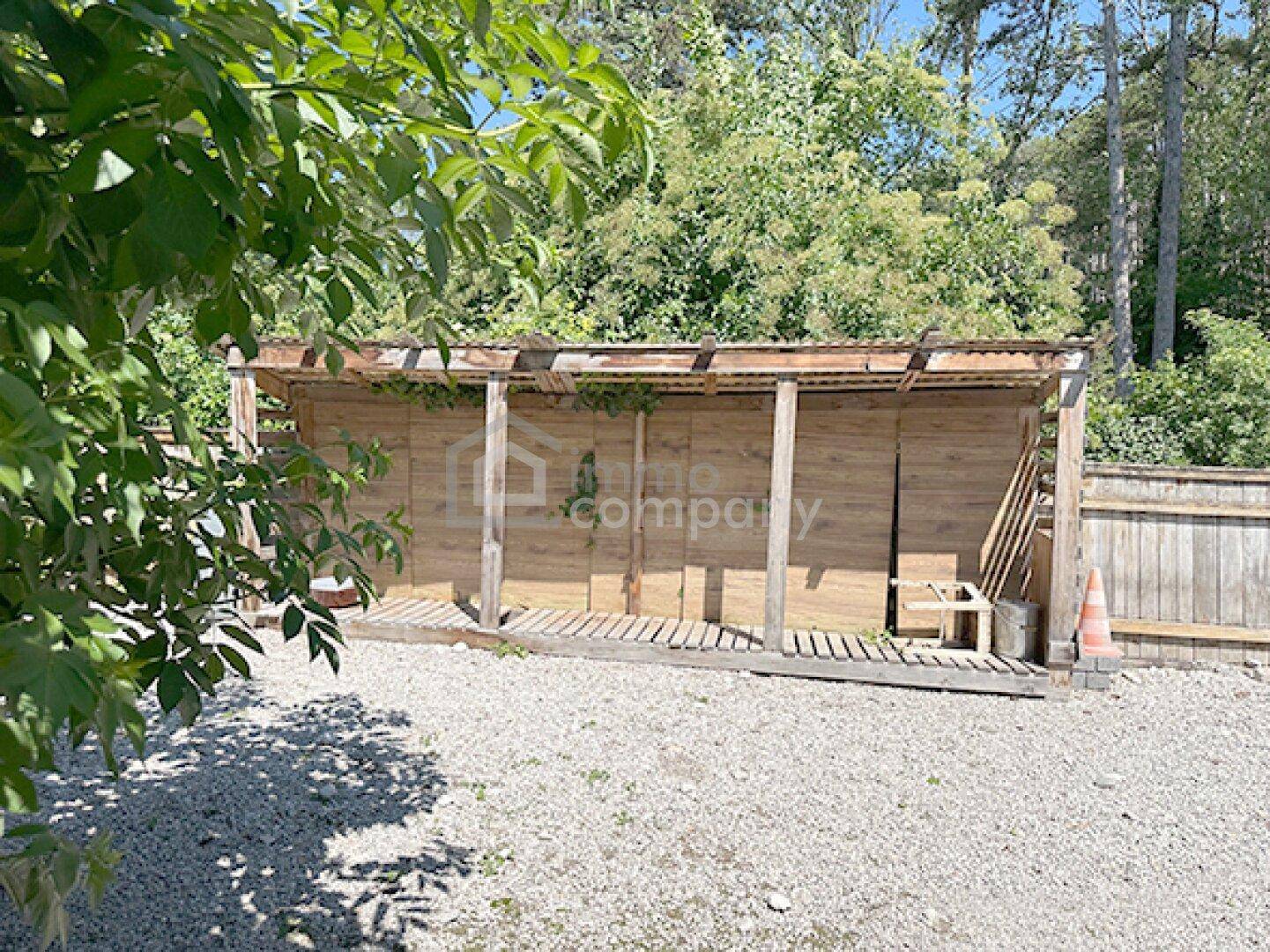 Offener Holzschuppen oder Carport mit Holzwänden und Kiesboden, umgeben von Bäumen.