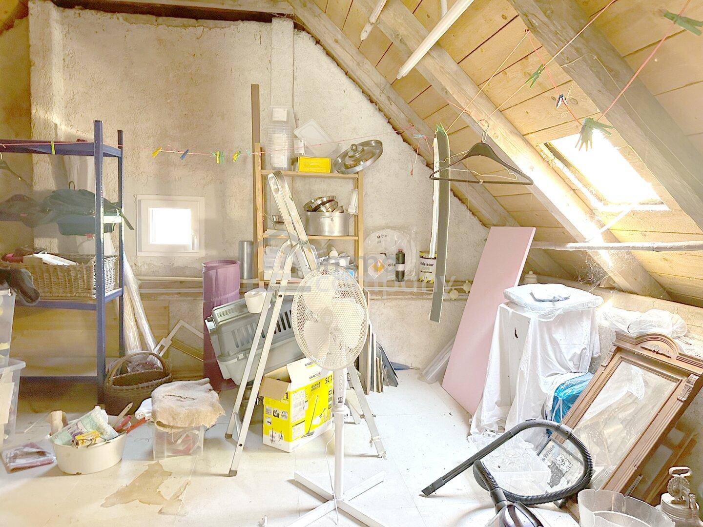 Unausgebauter Dachboden mit sichtbaren Holzbalken, einem Dachfenster und verschiedenen Lagergegenständen.