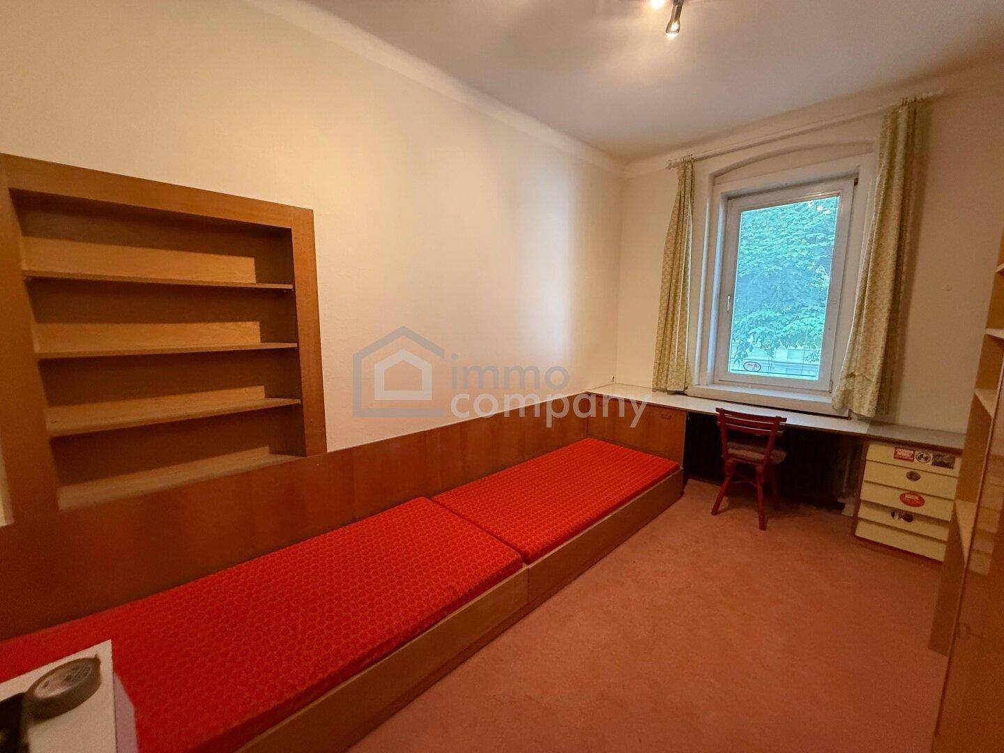 Schlafzimmer mit rotem Bett, integrierten Holzregalen und Fenster.