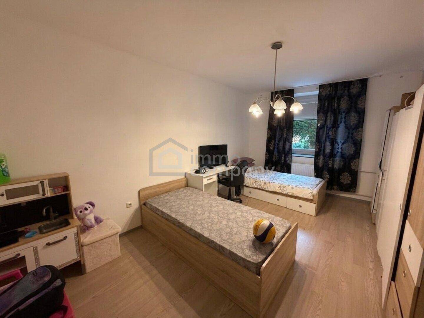 Kinderzimmer mit zwei Einzelbetten, Holzfußboden und einem Fenster mit dunklen Vorhängen.