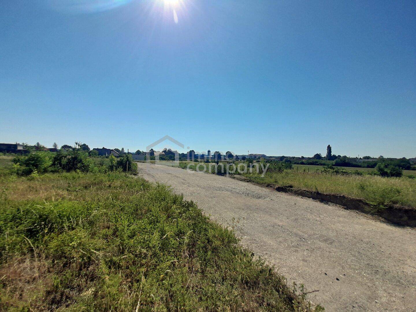 Unbefestigter Weg durch eine grüne Landschaft mit entfernten Gebäuden unter strahlendem Sonnenschein.