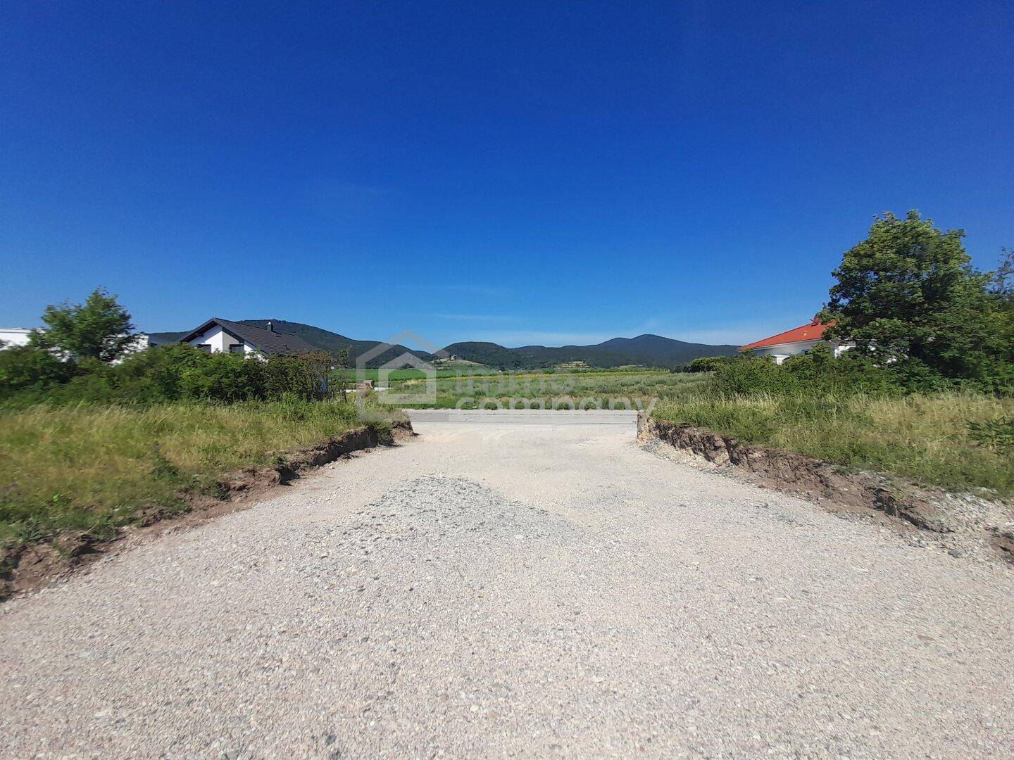 Schotterweg führt zu einer Betonkonstruktion auf einem Grundstück mit Häusern und Bergen im Hintergrund.