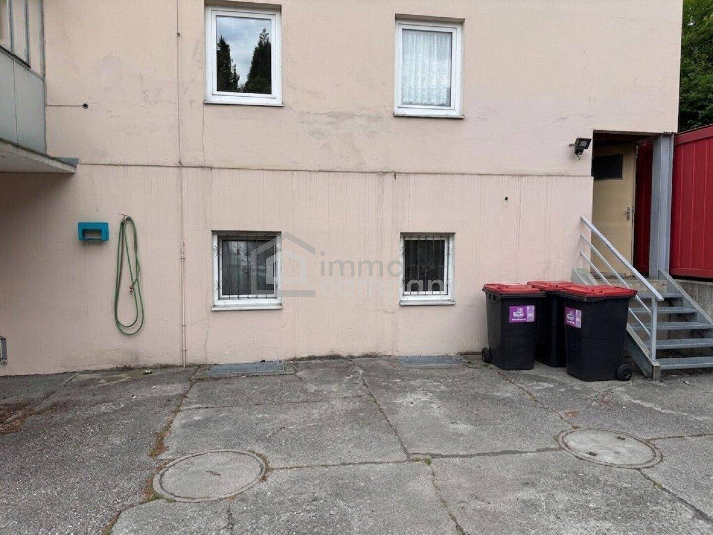 Außenansicht des Gebäudes mit Fenstern, Mülltonnen und einem Zugang über eine Treppe.