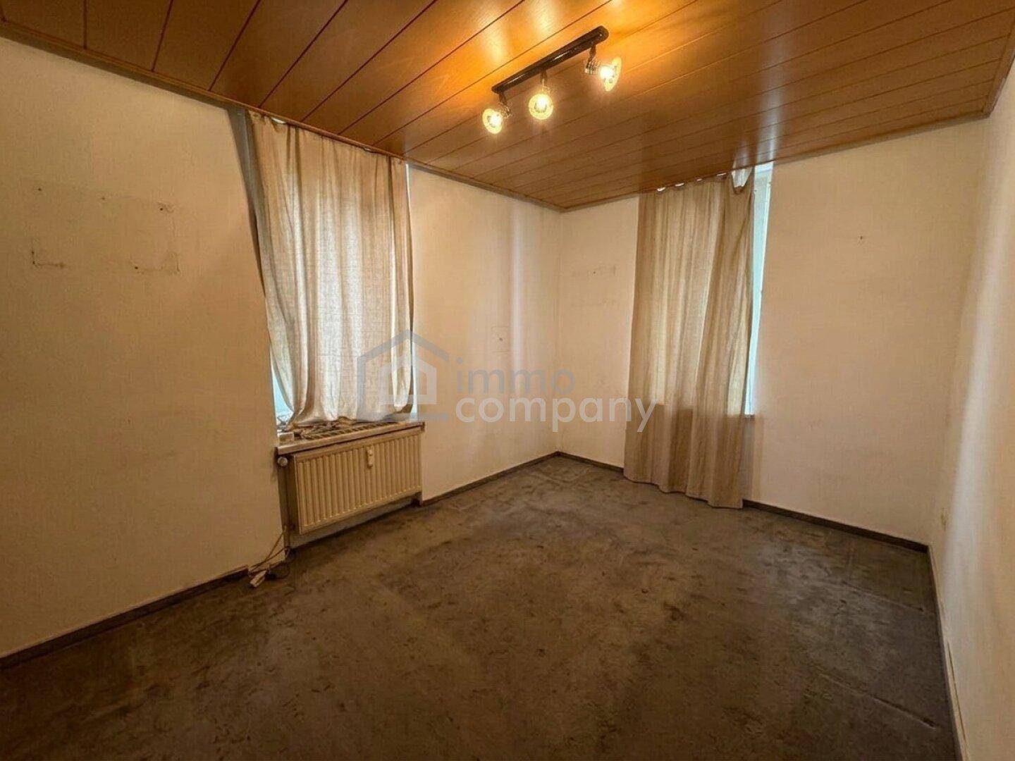 Leerer Raum mit dunklem Boden, Holzdecke und zwei Fenstern, renovierungsbedürftig.