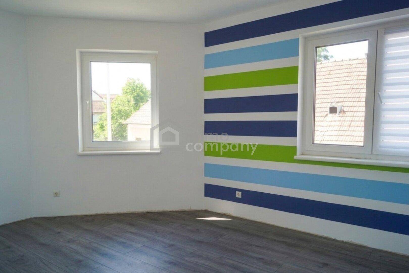 Helles Zimmer mit gestreifter Akzentwand, modernem Boden und zwei Fenstern.