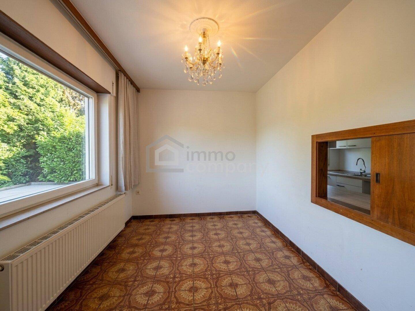 Leerer Raum mit Terrakotta-Fliesenboden, großem Fenster und Durchreiche zur Küche.