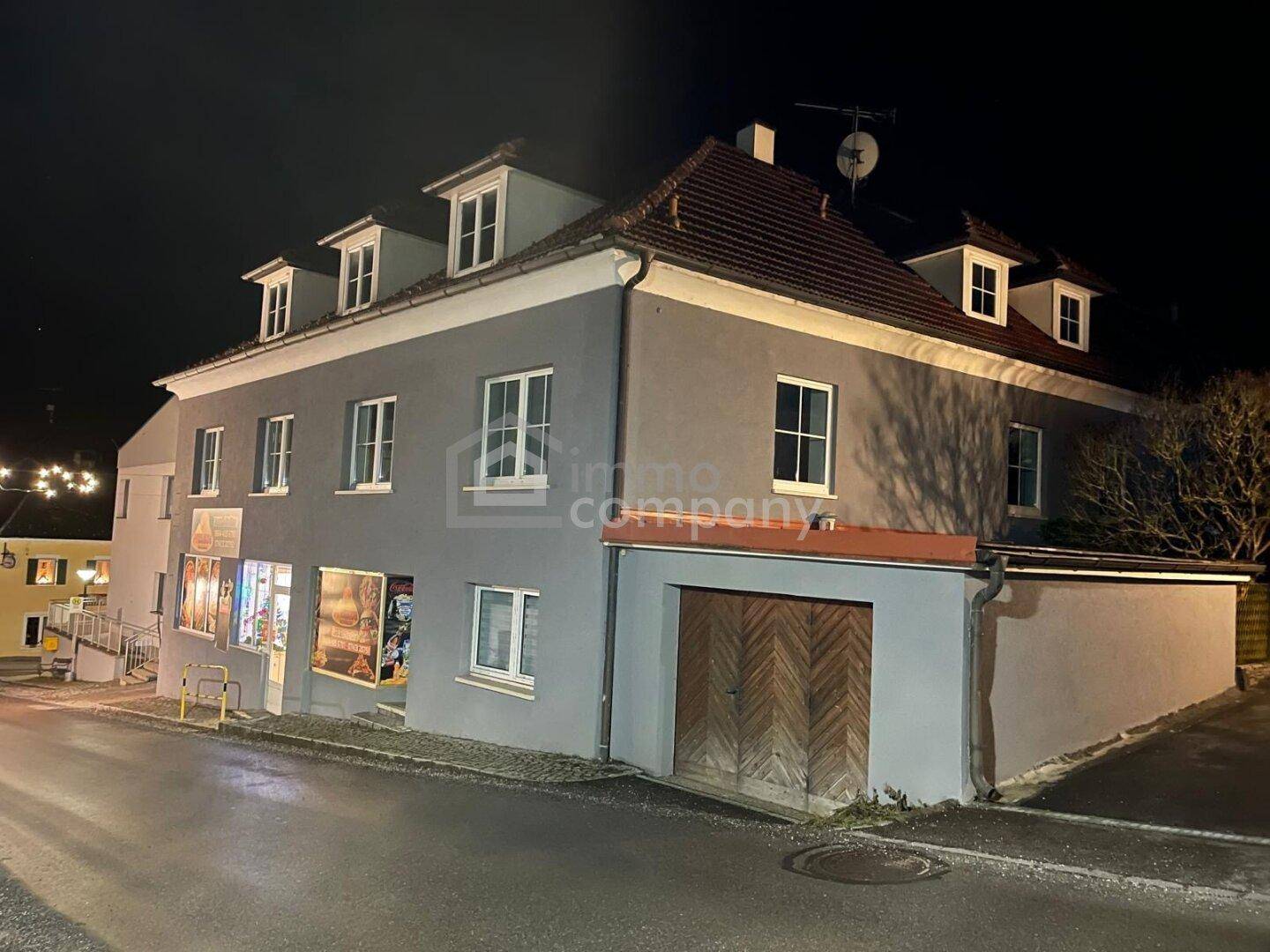Gebäudeansicht bei Nacht mit grauer Fassade, Dachgauben und einem Geschäft im Erdgeschoss.