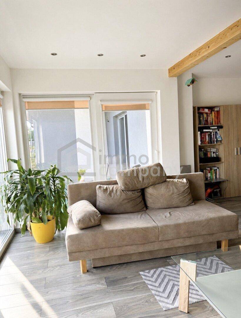 Heller Wohnbereich mit beigem Sofa, Bücherregal und viel Tageslicht durch große Fenster.