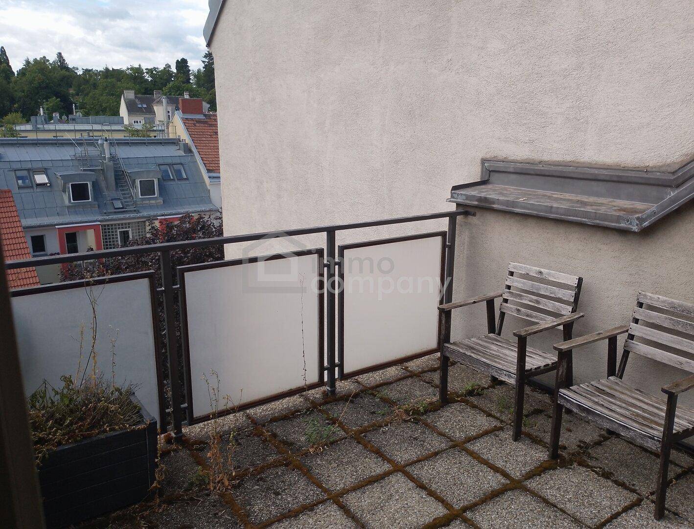 Kleine Dachterrasse mit Fliesenboden und Holzstühlen, umgeben von Stadtansichten und Dächern.