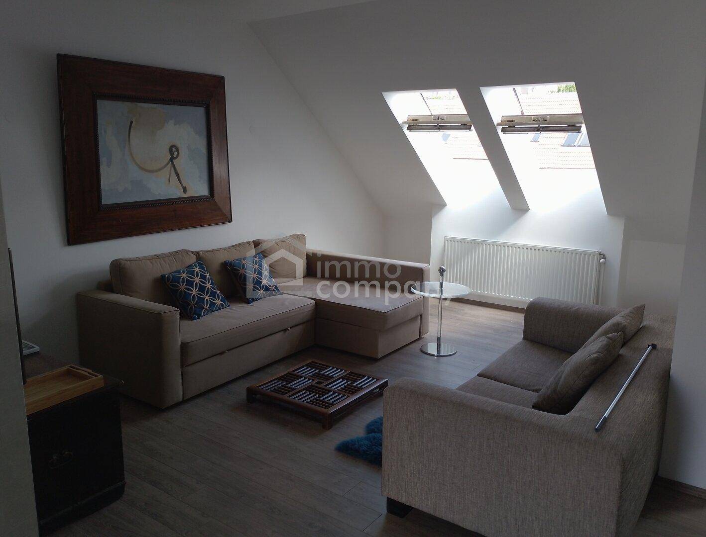 Gemütliches Wohnzimmer im Dachgeschoss mit beigem Sofa, Couchtisch und zwei großen Dachfenstern.