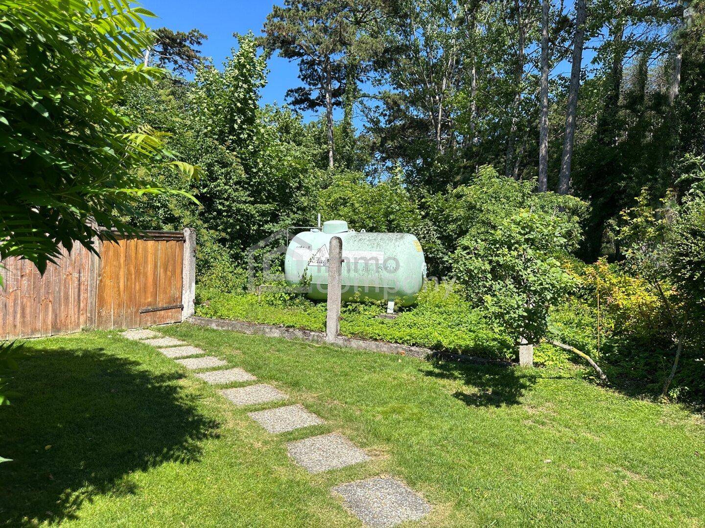 Großer Gastank im Garten, umgeben von Bäumen und einem gepflegten Rasenweg.