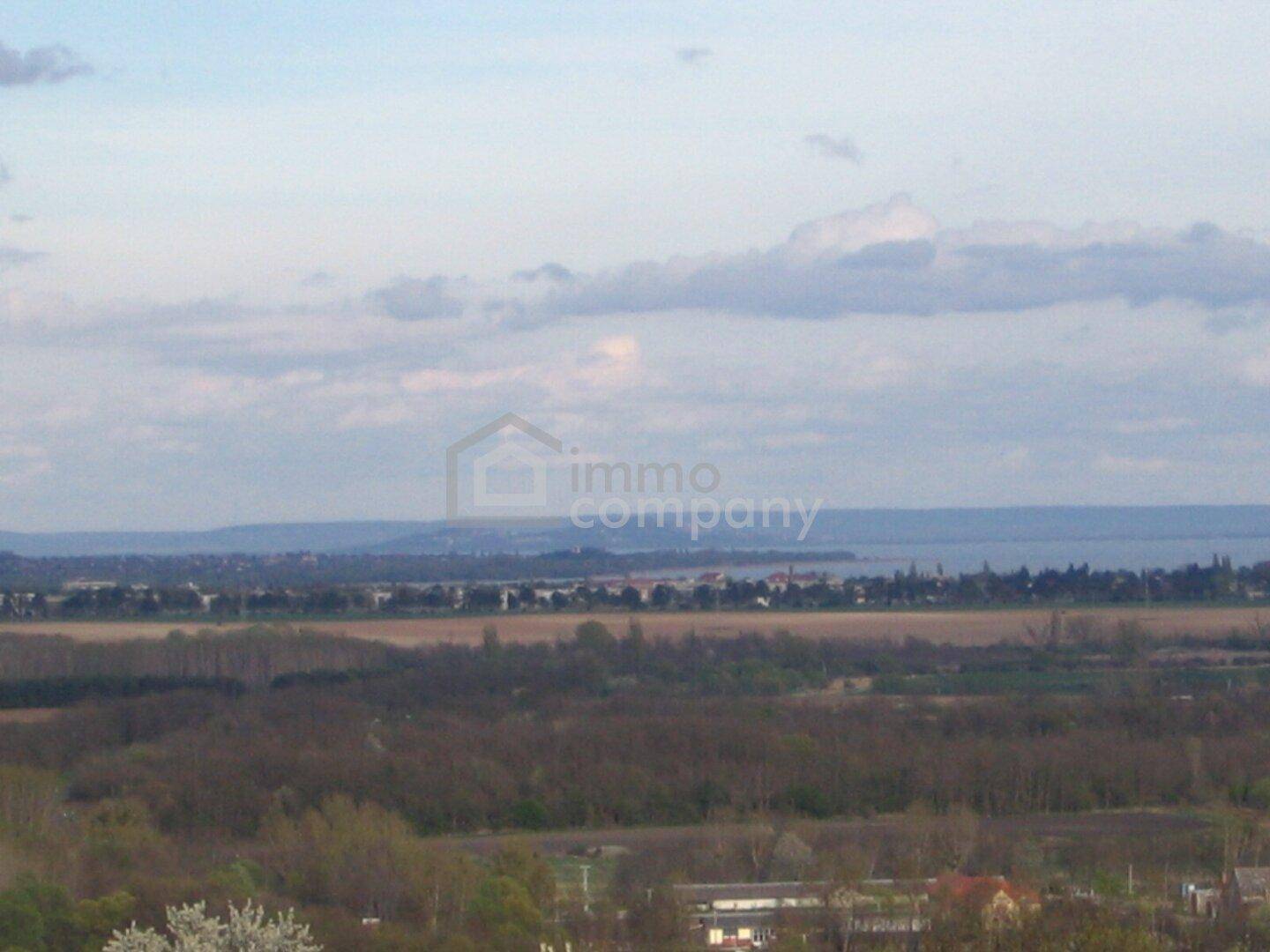 Weitläufiger Blick über eine Landschaft mit Feldern und einem großen See am Horizont unter bewölktem Himmel.