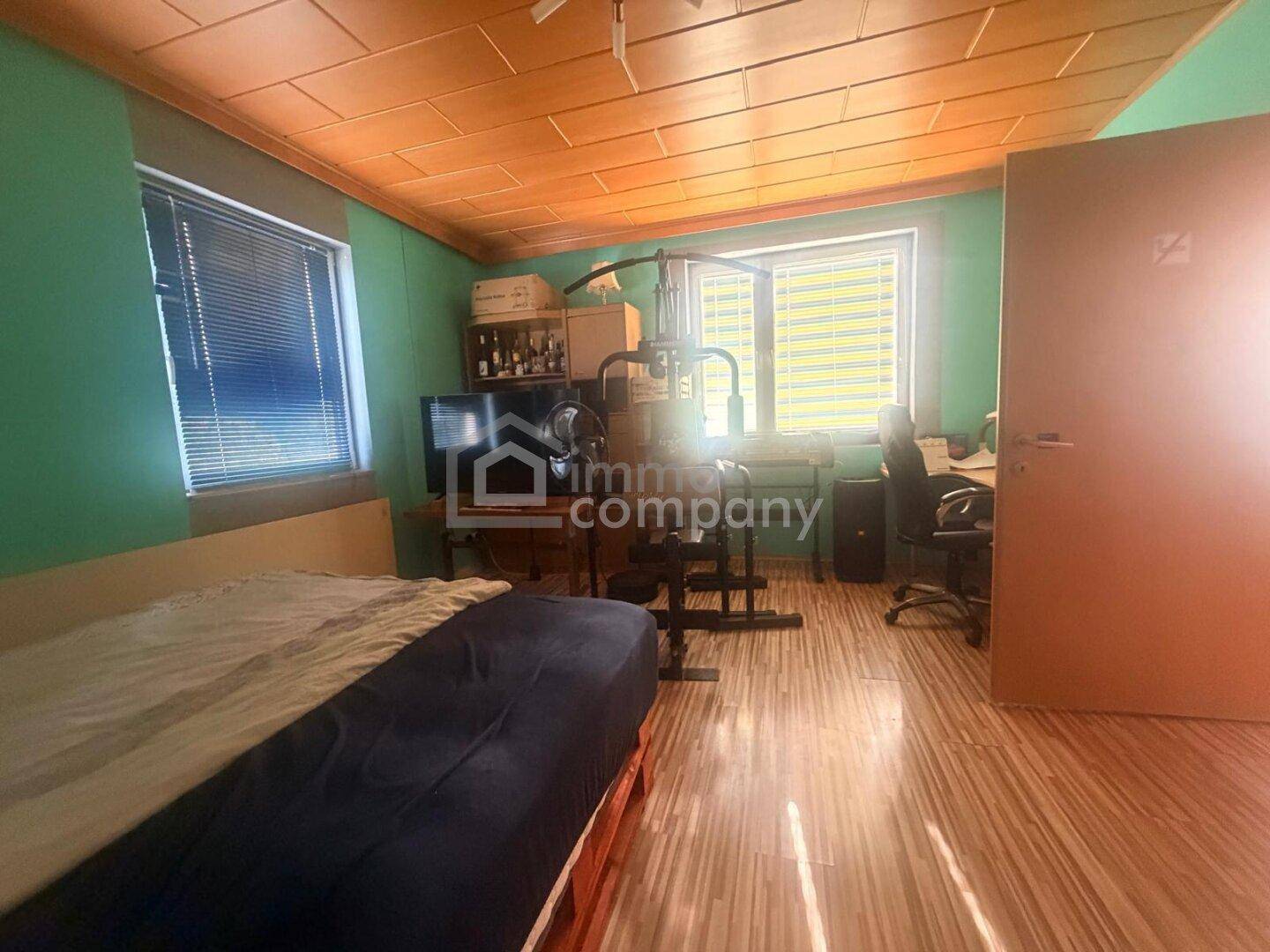 Vielseitiges Zimmer mit Bett, Schreibtisch und Fitnessgeräten, hell durch große Fenster.