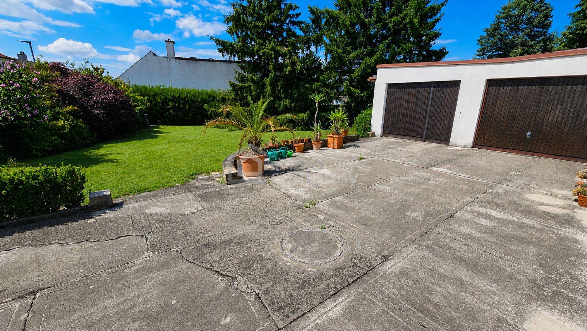 Betonierte Einfahrt mit Doppelgarage und angrenzendem grünen Gartenbereich unter blauem Himmel.