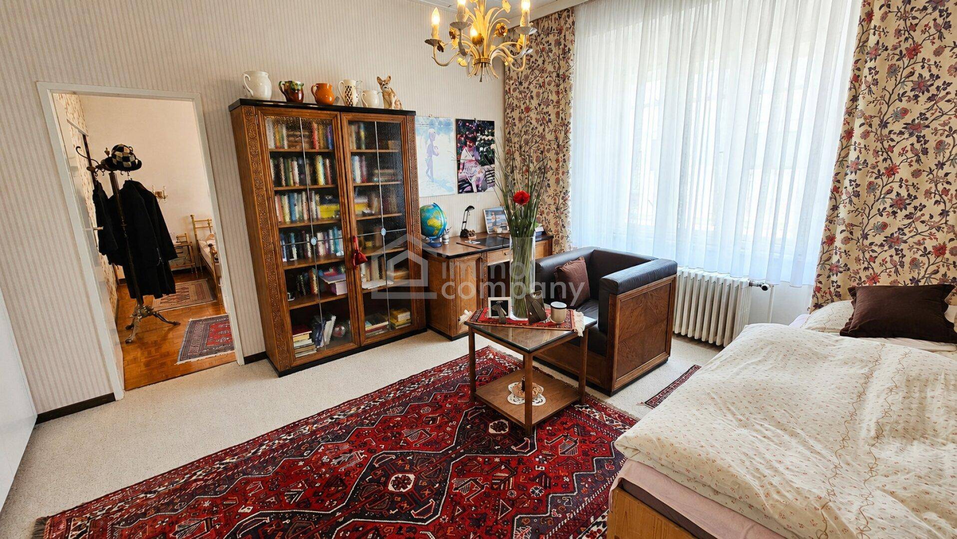 Schlafzimmer mit Teppichboden, Bücherregal und Schreibtisch, hell durch große Fenster.