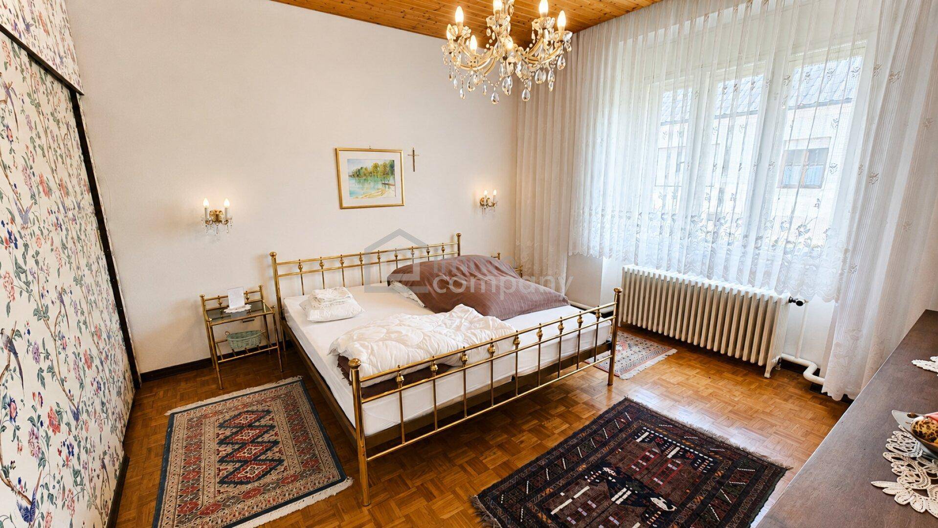 Schlafzimmer mit Parkettboden, Messingbett und Kronleuchter, hell durch große Fenster.