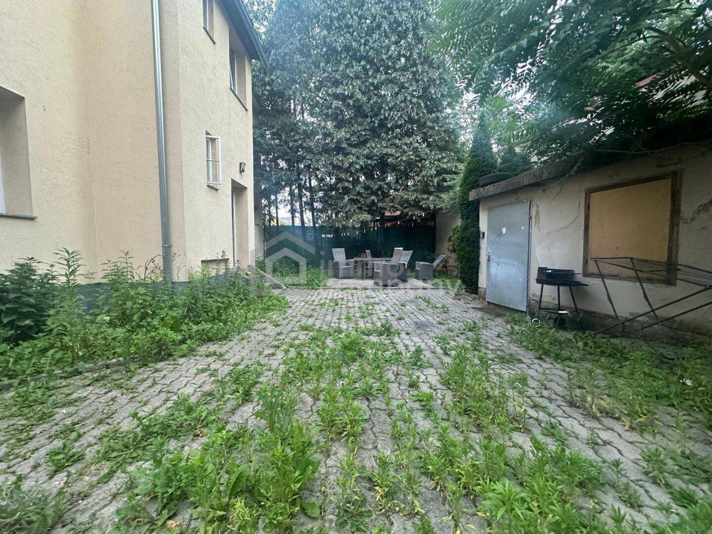 Gepflasterter Außenbereich mit Sitzgelegenheiten, umgeben von viel Grün und einem Garagengebäude.