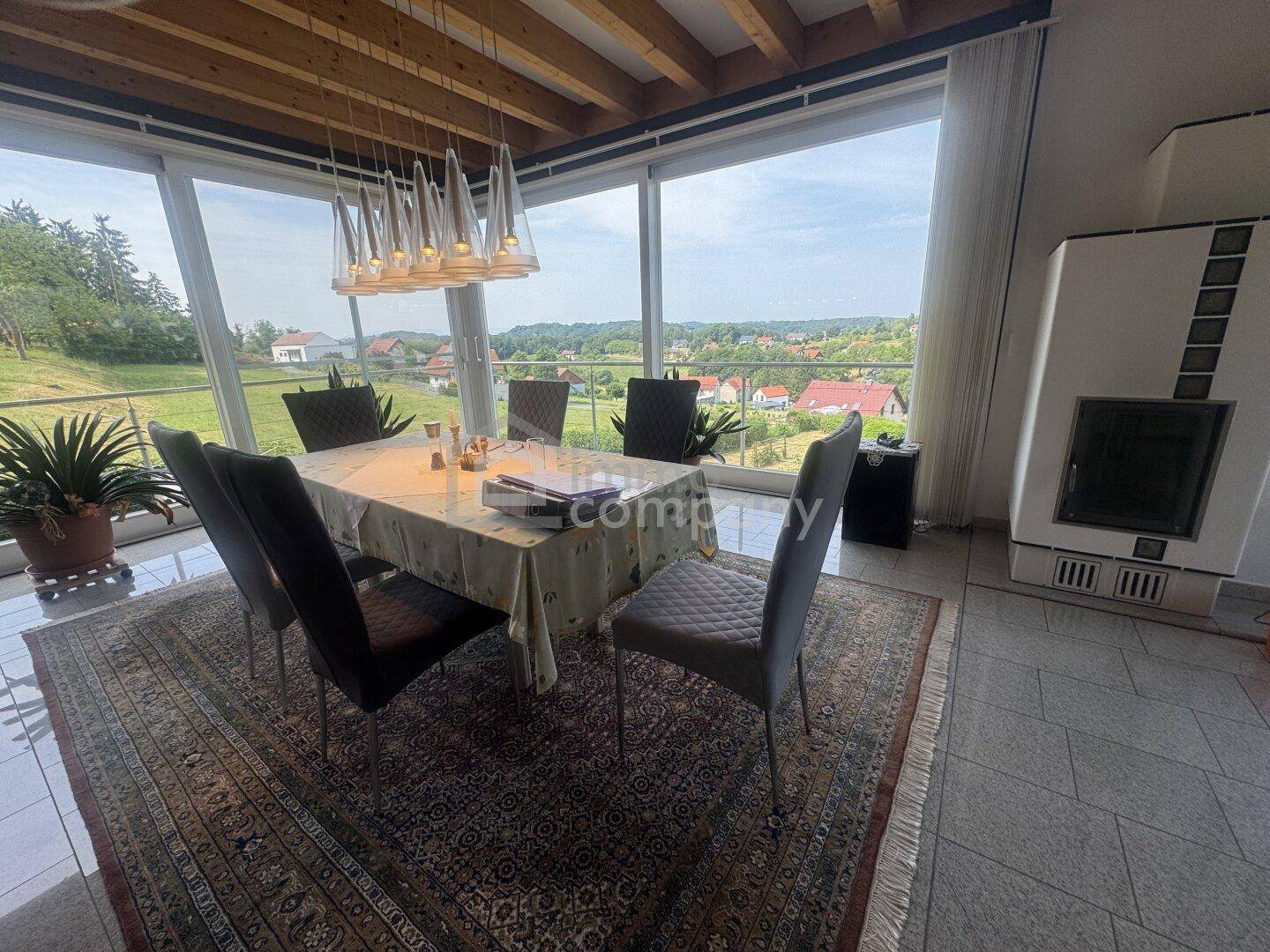 Essbereich mit großem Tisch, modernen Stühlen und Kachelofen, umgeben von Panoramafenstern.