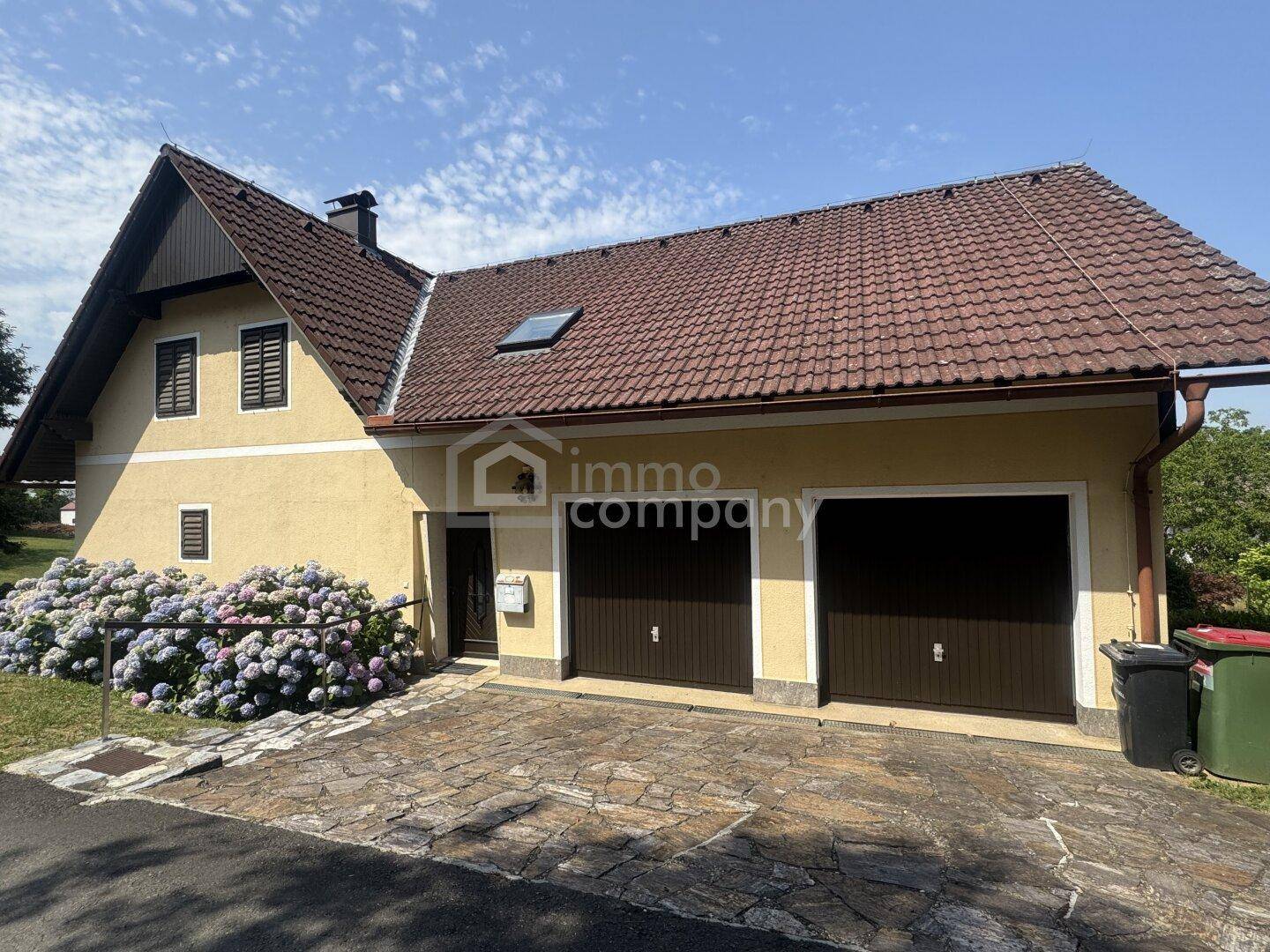 Vorderansicht des Hauses mit Doppelgarage, gepflasterter Einfahrt und blühenden Hortensien.