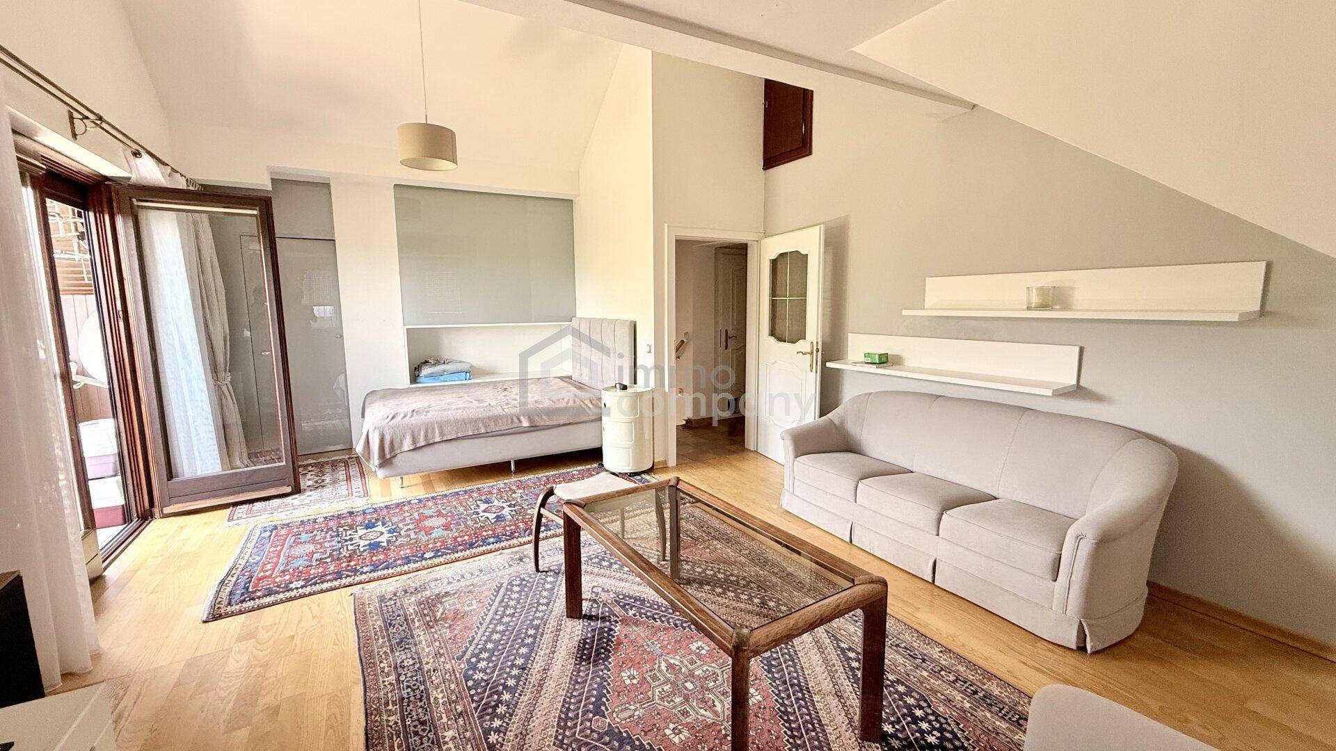 Helles Schlafzimmer mit Bett, Sofa und Zugang zu einem Balkon, unter einem Dachschrägenbereich.