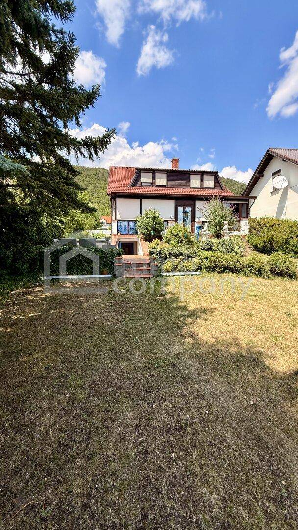 Hausfassade mit rotem Dach, umgeben von einem Garten und Bäumen, mit Hügeln im Hintergrund.