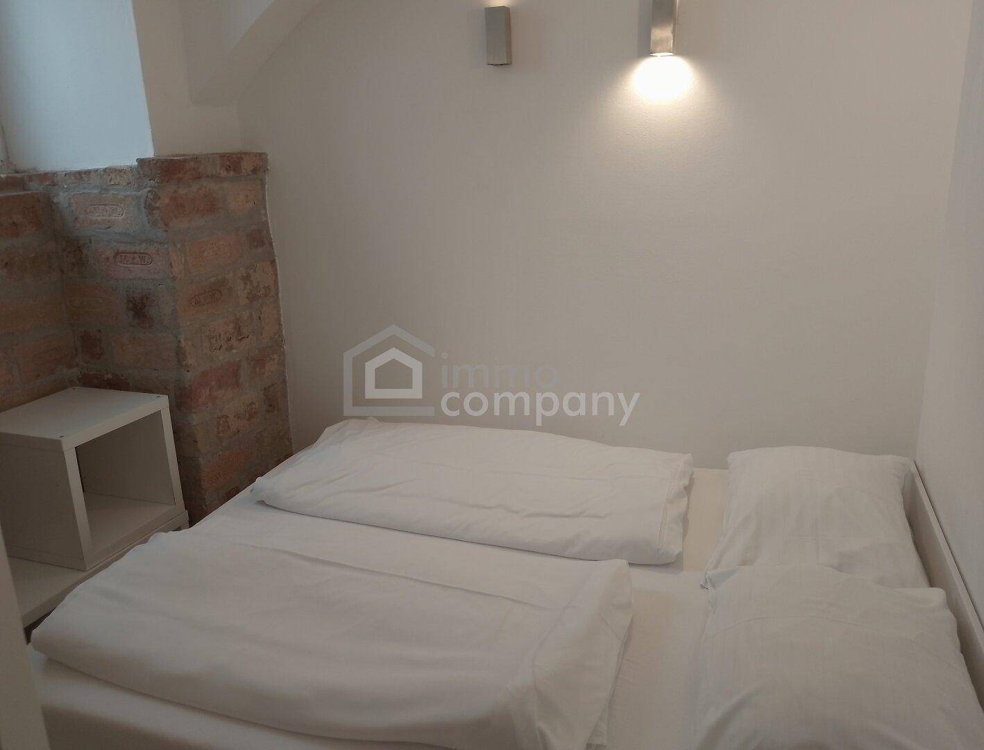 Schlafzimmer mit Doppelbett, weißer Bettwäsche und einem kleinen Nachttisch neben der Ziegelwand.