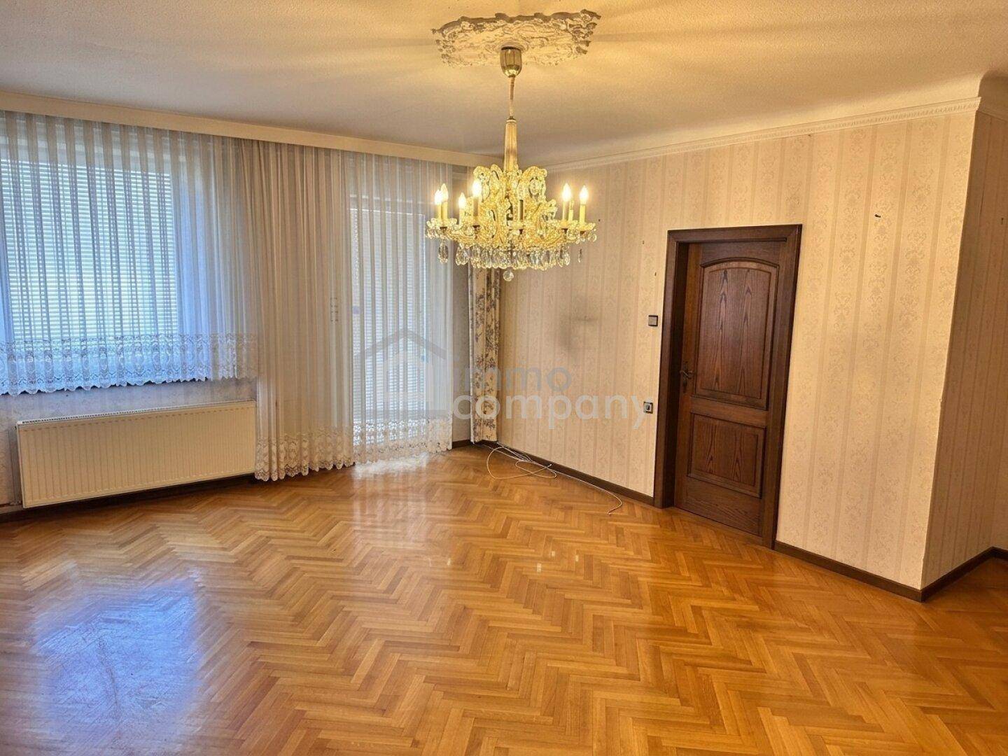 Helles Wohnzimmer mit Fischgrätparkett, gemusterter Tapete und klassischem Kronleuchter.
