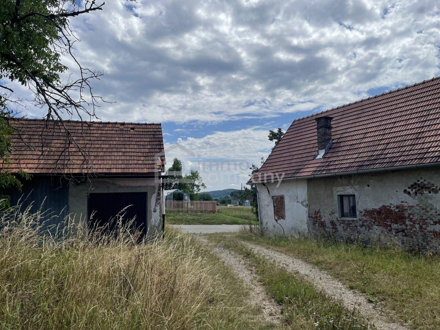 Zwei alte Gebäude mit Ziegeldächern und einem Feldweg unter bewölktem Himmel.