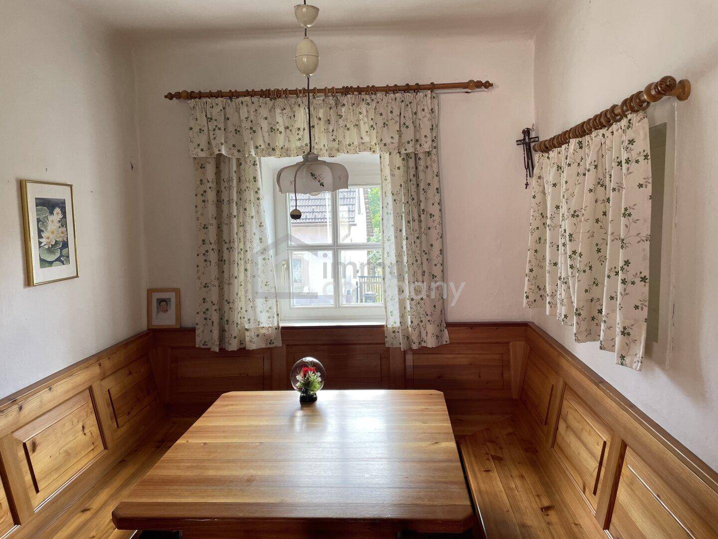 Gemütlicher Essbereich mit Holztisch, Eckbank und floralen Vorhängen unter einem Fenster.
