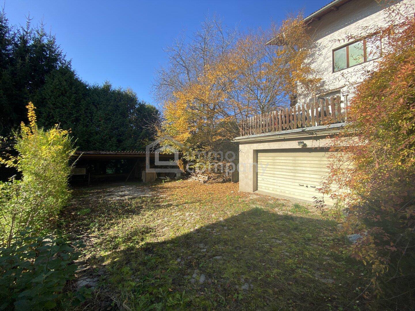 Gartenbereich mit Garage und Carport, umgeben von herbstlicher Vegetation unter blauem Himmel.