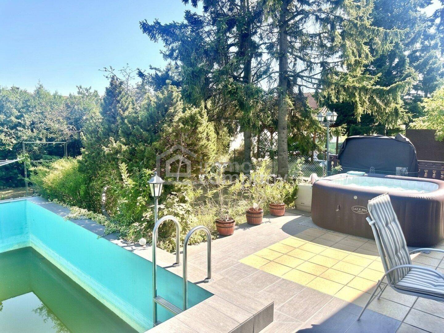 Sonniger Terrassenbereich mit Pool, Whirlpool und Sitzgelegenheiten, umgeben von hohen Bäumen.
