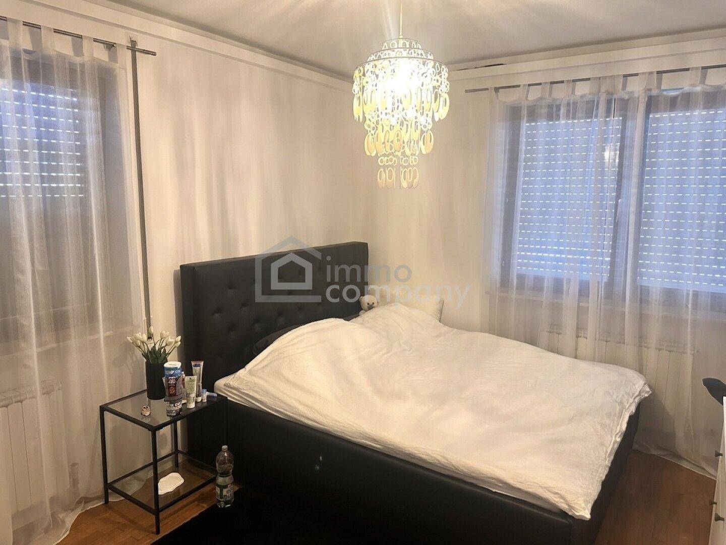Schlafzimmer mit Doppelbett, Nachttisch und Kronleuchter, beleuchtet durch zwei Fenster.