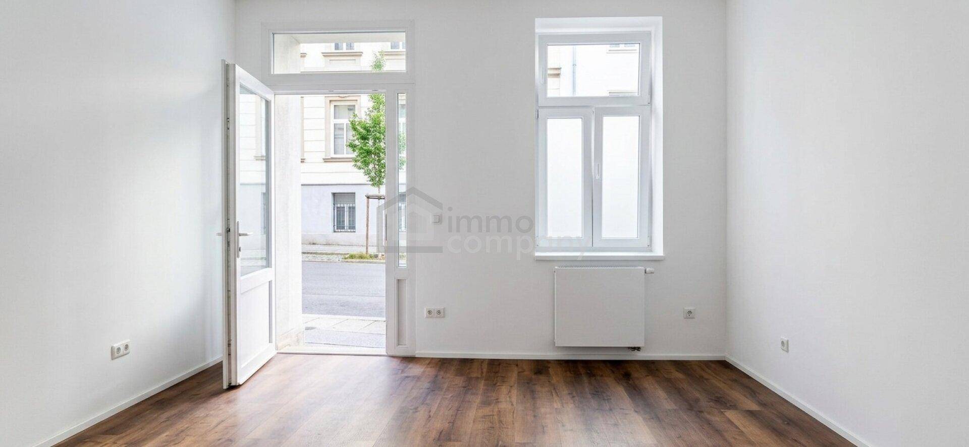 Geräumiger, leerer Raum mit dunklem Laminatboden, weißen Wänden und einem Heizkörper unter dem Fenster.