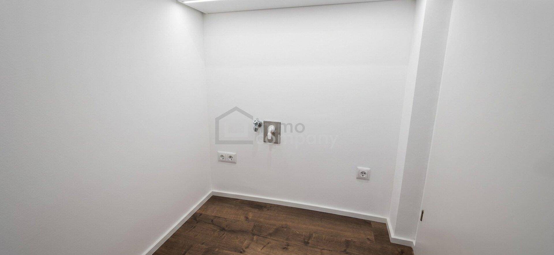 Kleiner, leerer Raum mit weißen Wänden, dunklem Holzboden und sichtbaren Anschlüssen für Waschmaschine oder Trockner.