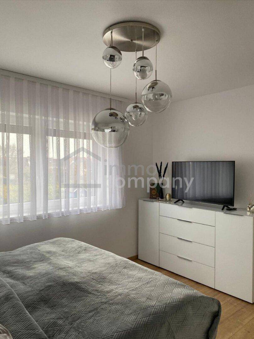 Schlafzimmer mit großem Fenster, modernem Deckenlicht und einem Sideboard unter dem Fernseher.