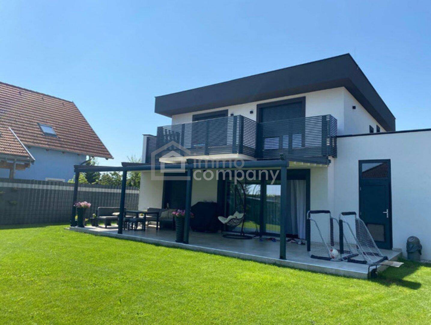 Rückseite des modernen Hauses mit großer Rasenfläche und überdachter Terrasse für Mahlzeiten im Freien.