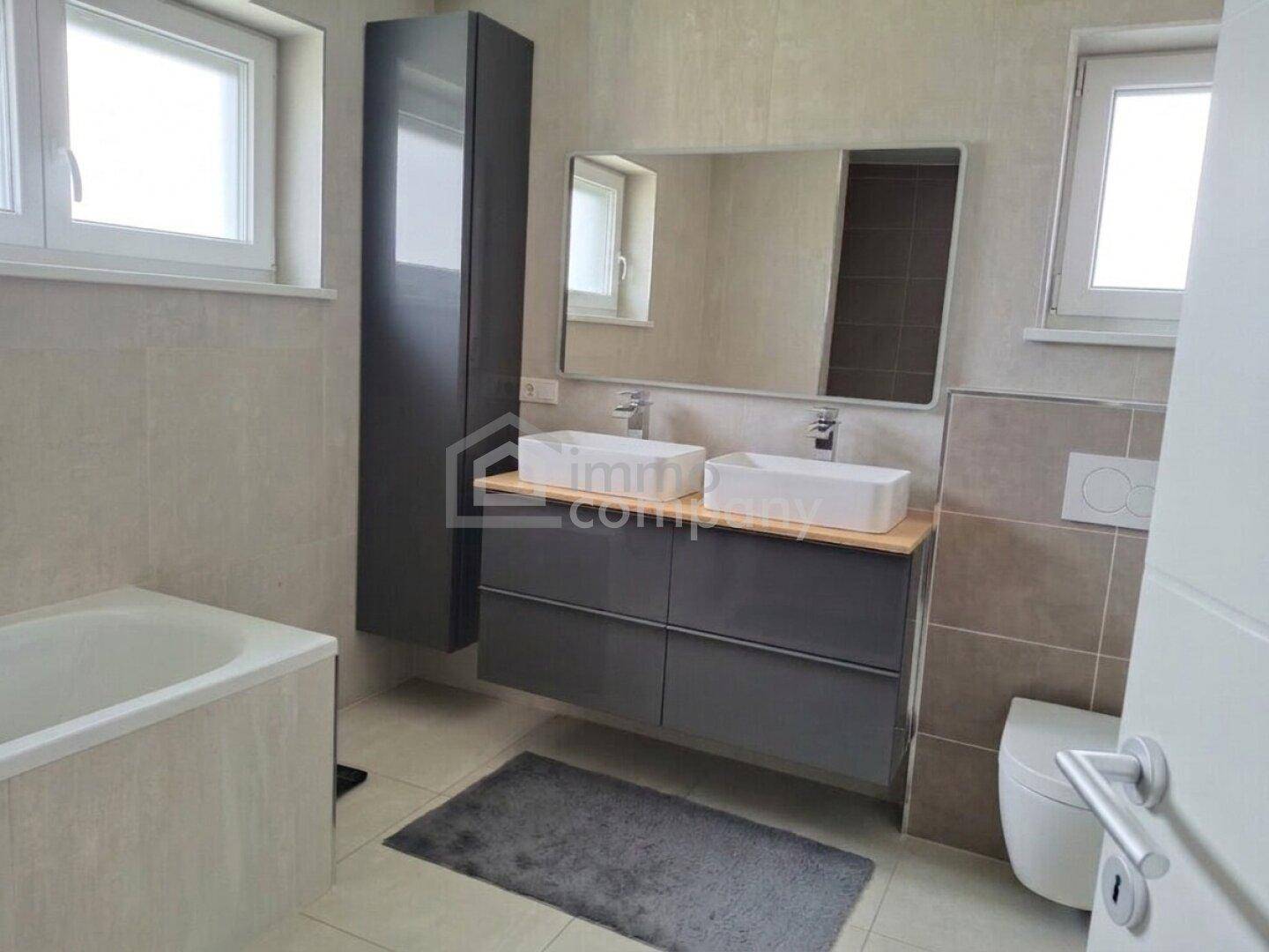 Modernes Badezimmer mit Doppelwaschbecken, Badewanne und zwei Fenstern für Tageslicht.