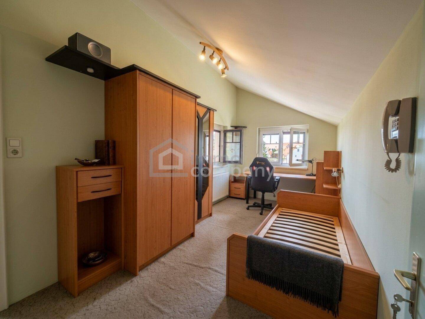 Dachzimmer mit schrägen Wänden, Teppichboden und Holzmöbeln, eingerichtet als Schlafzimmer.