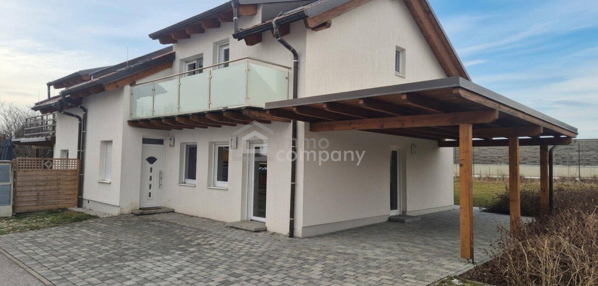 Modernes Einfamilienhaus mit weißer Fassade, Balkon mit Glasgeländer und einem Holzcarport.