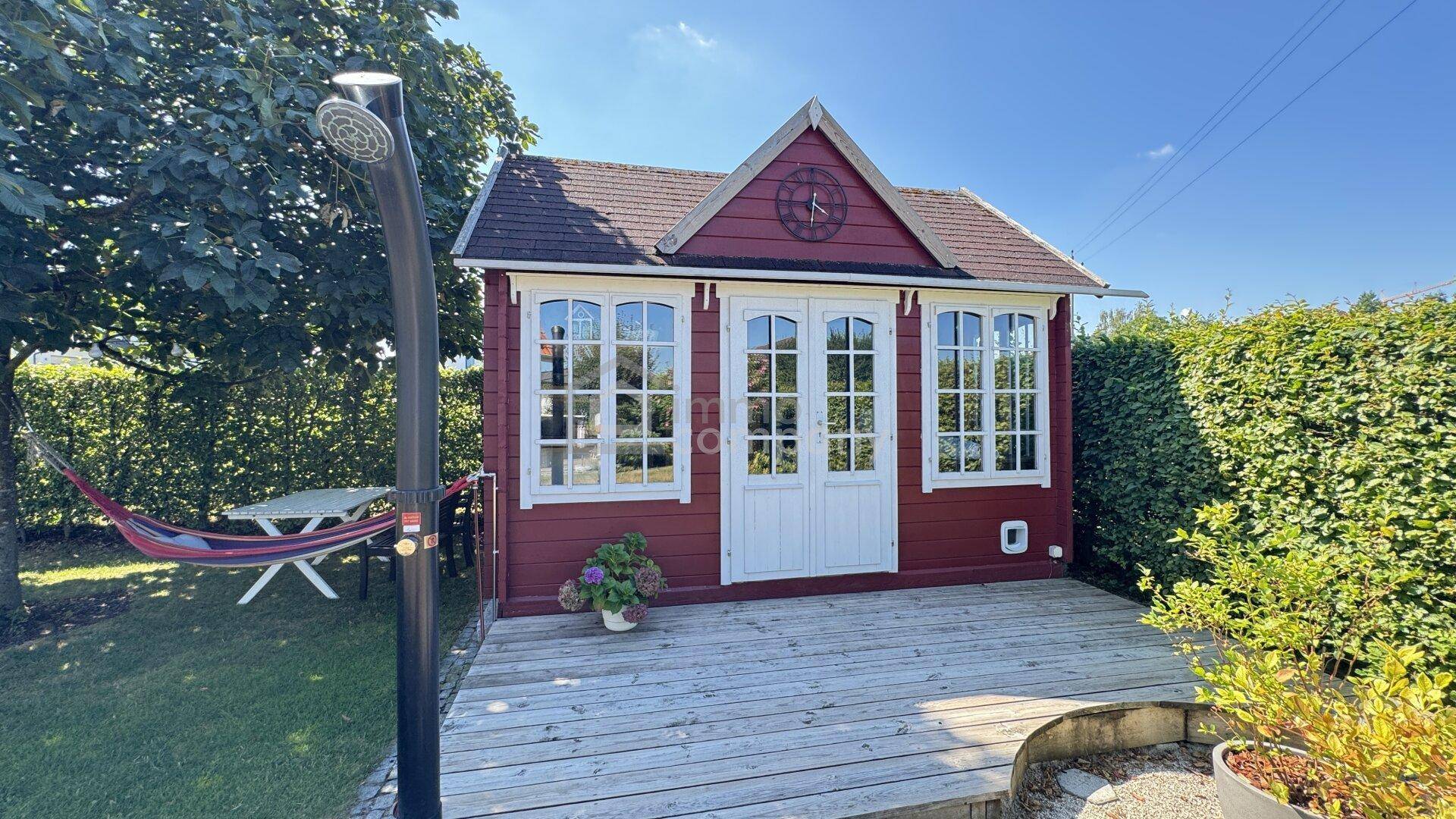 Rotes Gartenhaus mit Holzterrasse, Außendusche und Hängematte im grünen Garten.