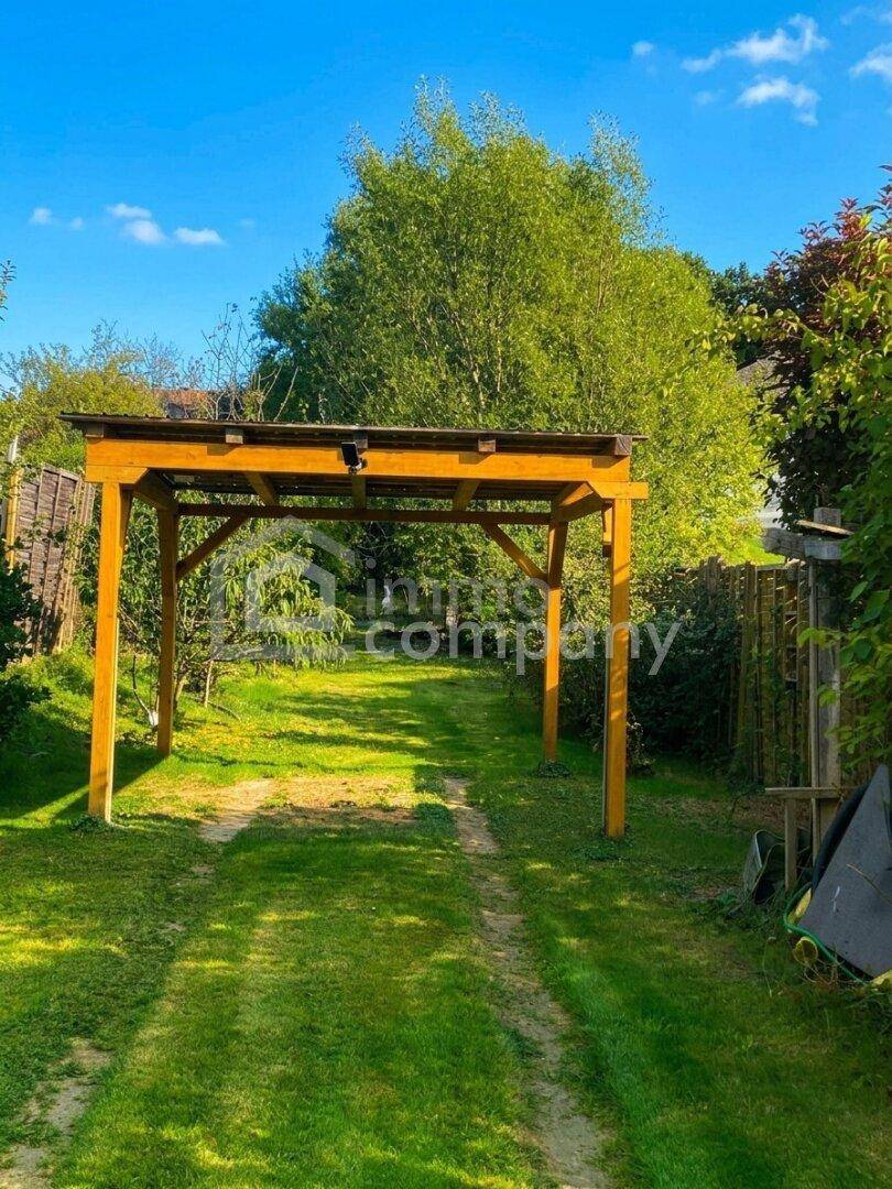 Hölzerner Carport über einer grasbewachsenen Einfahrt, umgeben von Bäumen und Zäunen.