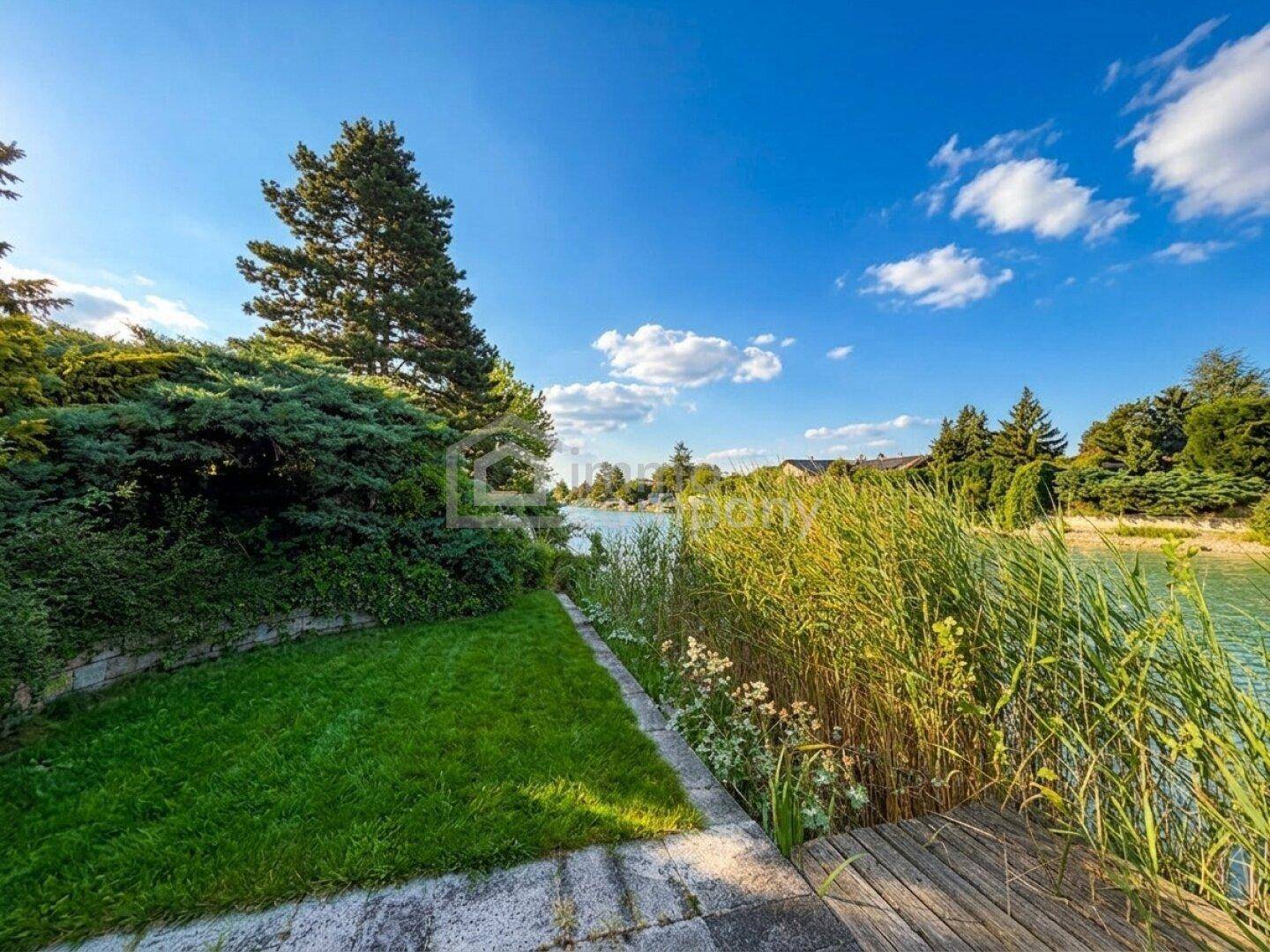 Uferbereich mit Schilf, kleiner Rasenfläche und Holzsteg, der zum ruhigen Wasser führt.