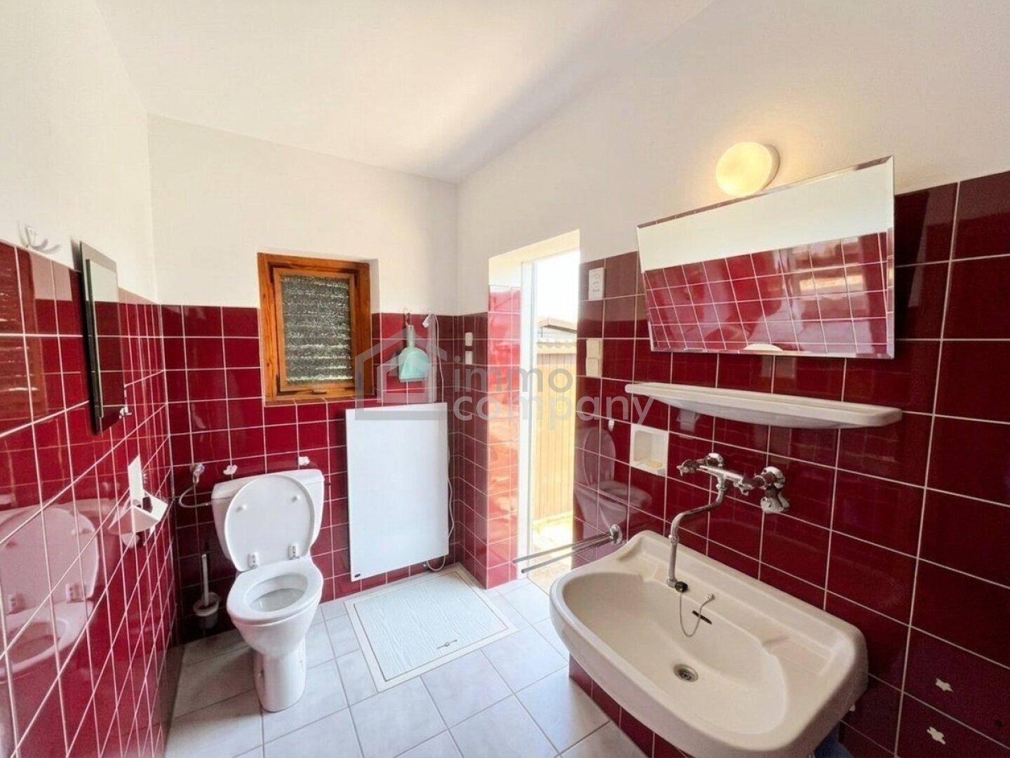 Badezimmer mit roten Fliesen, Toilette, Waschbecken und einem Fenster für natürliche Belüftung.